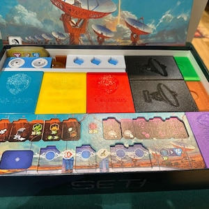 May include: A board game box with various colorful game pieces. The box contains blue, yellow, red, black, and green components, along with illustrated cards and a background image of satellite dishes. The word "SETI" is visible on the box.