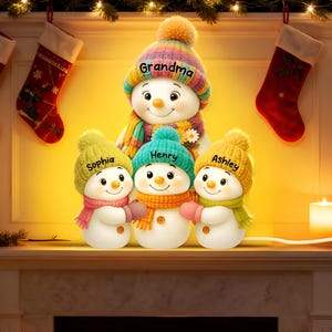 May include: A family of snowman figurines with personalized names on their hats: "Grandma", "Sophia", "Henry", and "Ashley". Each snowman wears a colorful hat, scarf, and mittens. The scene is set on a mantel with Christmas stockings and a lit candle.