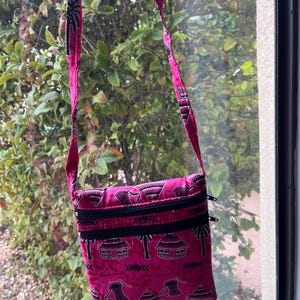 May include: A pink crossbody bag with a black zipper and a black and white print of houses and palm trees.