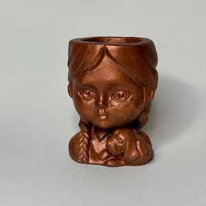 Könnte beinhalten: Ein kupferfarbener Keramik-Pflanztopf in Form eines Mädchenkopfes mit einem kleinen Hund in ihren Armen. Der Topf hat eine strukturierte Oberfläche und eine kleine Öffnung oben zum Pflanzen.
