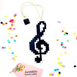 Puede incluir: Adorno de clave de sol en pixel art en blanco y negro con una cuerda dorada. El adorno está rodeado de cuentas de plástico de colores.