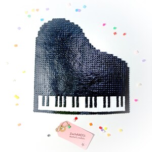 Può includere: Un pianoforte a coda nero realizzato con perline di plastica con tasti bianchi. Il pianoforte è su una superficie bianca con perline colorate sparse intorno. Un'etichetta rosa con testo si trova nell'angolo in basso a destra.