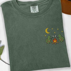 Könnte beinhalten: Ein grünes T-Shirt mit Rundhalsausschnitt und kurzen Ärmeln. Das Shirt hat ein gesticktes Design von zwei Schildkröten, die Marshmallows über einem Lagerfeuer unter einem Mond und Sternen rösten. Das Shirt ist aus einem weichen, bequemen Material.