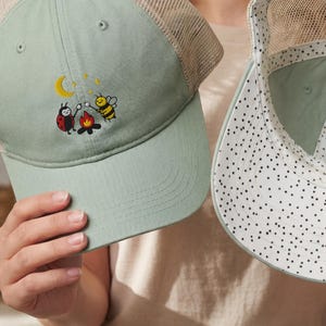 May include: Two sage green trucker caps with beige mesh backs. One cap features an embroidered graphic of a ladybug and a bee roasting marshmallows over a campfire under a crescent moon. The other cap shows the interior with a black polka dot lining.