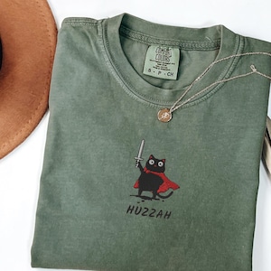 Può includere: Una maglietta verde muschio con un gatto nero dei cartoni animati con un mantello rosso e una spada, con la parola "Huzzah" sotto. La maglietta ha un girocollo e maniche corte. Una collana d'oro è drappeggiata sulla maglietta.