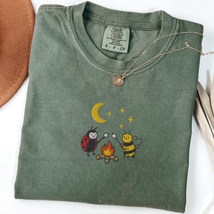 May include: An olive green short-sleeve t-shirt featuring an embroidered graphic of a ladybird and a bee roasting marshmallows over a campfire under a yellow crescent moon and stars. The inner neck label reads Comfort Colors S P CH.