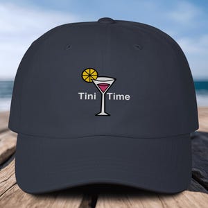 May include: Navy blue baseball cap featuring an embroidered cocktail glass with a lemon slice and the words "Tini Time." The cap has a curved brim and a classic six-panel design. The background shows a beach scene.