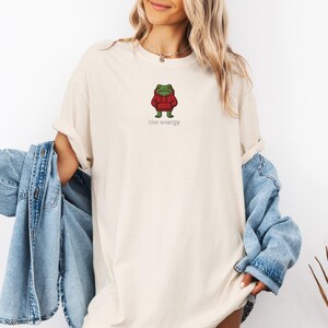 May include: Cream-colored t-shirt featuring a cartoon frog wearing a red hoodie. The frog is standing with its arms at its sides. The words "low energy" are printed in a small, gray font below the frog. The shirt is a relaxed fit.