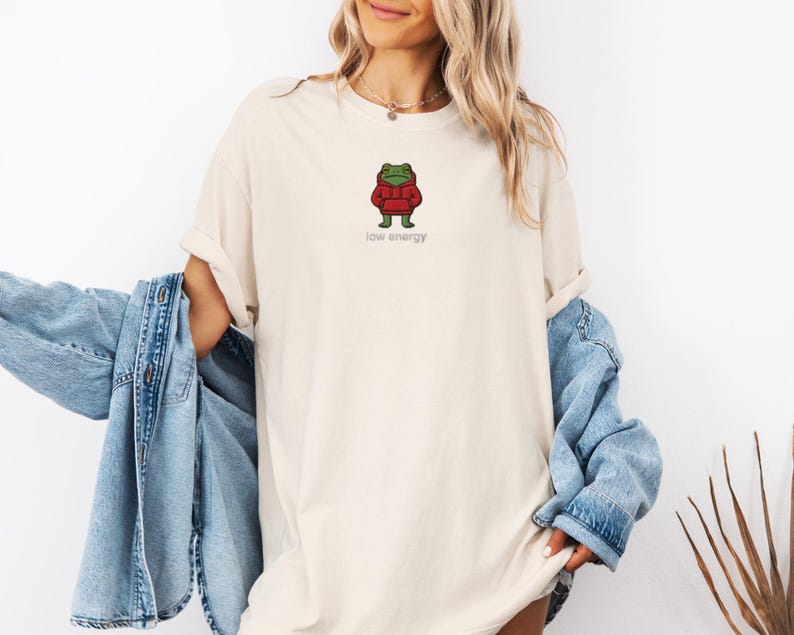 May include: Cream-colored t-shirt featuring a cartoon frog wearing a red hoodie. The frog is standing with its arms at its sides. The words "low energy" are printed in a small, gray font below the frog. The shirt is a relaxed fit.
