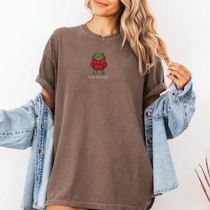May include: Brown t-shirt with a cartoon frog wearing a red hoodie. The text "low energy" is below the frog. The shirt is worn with a blue denim jacket.