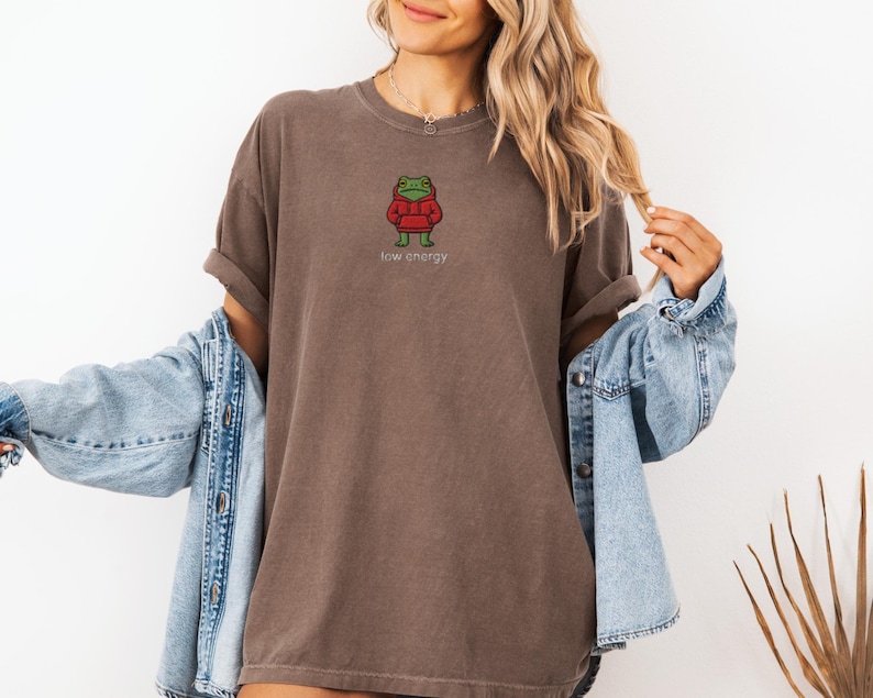 May include: Brown t-shirt with a cartoon frog wearing a red hoodie. The text "low energy" is below the frog. The shirt is worn with a blue denim jacket.