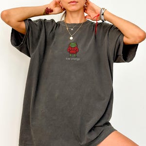 May include: Dark grey oversized t-shirt featuring a cartoon frog wearing a red sweater. The frog is embroidered on the front with the words "low energy" below it. The shirt has short sleeves and a relaxed fit.
