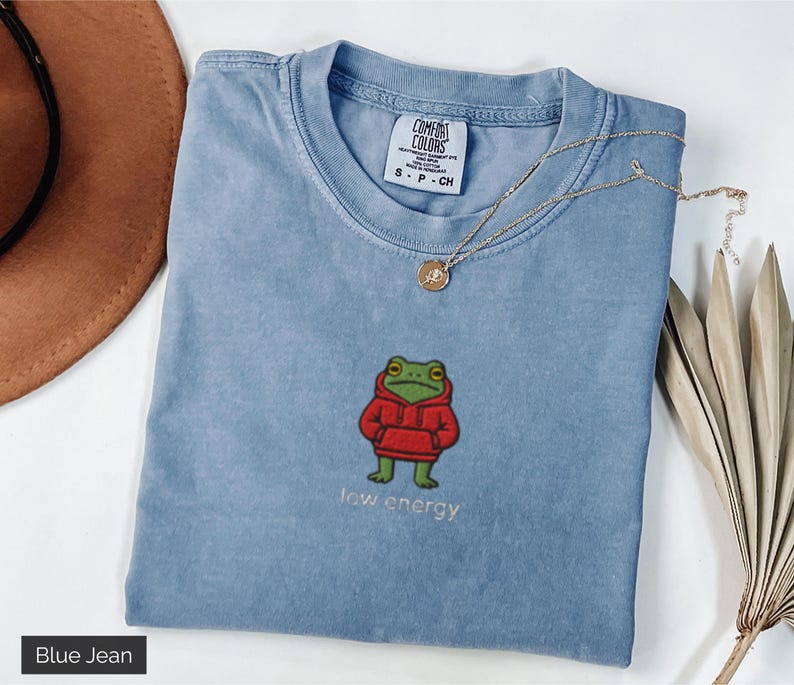 May include: A blue jean colored t-shirt with an embroidered frog wearing a red hoodie. The text "low energy" is embroidered below the frog. A gold necklace is draped across the shirt.