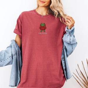 May include: A red-orange t-shirt featuring a cartoon frog wearing a red hoodie. The frog has green eyes and is embroidered on the front. The text "low energy" is below the frog. The shirt is worn with a blue denim jacket.