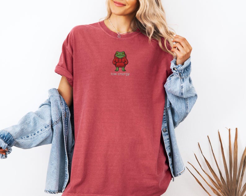 May include: A red-orange t-shirt featuring a cartoon frog wearing a red hoodie. The frog has green eyes and is embroidered on the front. The text "low energy" is below the frog. The shirt is worn with a blue denim jacket.