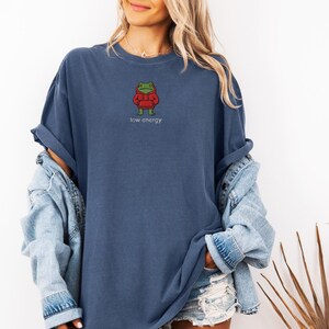 May include: A navy blue t-shirt with a small embroidered frog wearing a red hoodie. The text "low energy" is printed below the frog. The shirt is a relaxed fit.