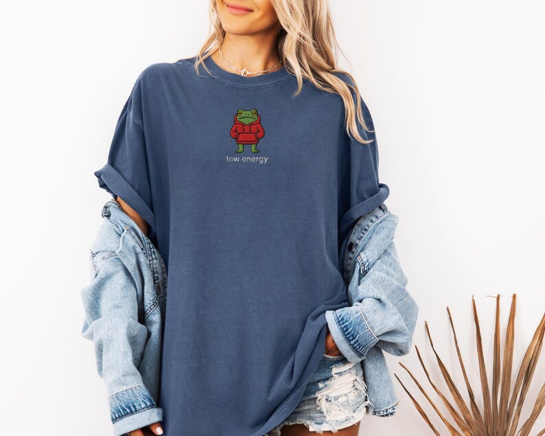 May include: A navy blue t-shirt with a small embroidered frog wearing a red hoodie. The text "low energy" is printed below the frog. The shirt is a relaxed fit.