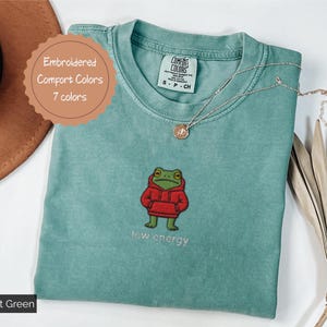 May include: Light green t-shirt with an embroidered frog wearing a red hoodie. The text "low energy" is below the frog. The shirt has a Comfort Colors tag and a round tag that says "Embroidered Comfort Colors 7 colors".