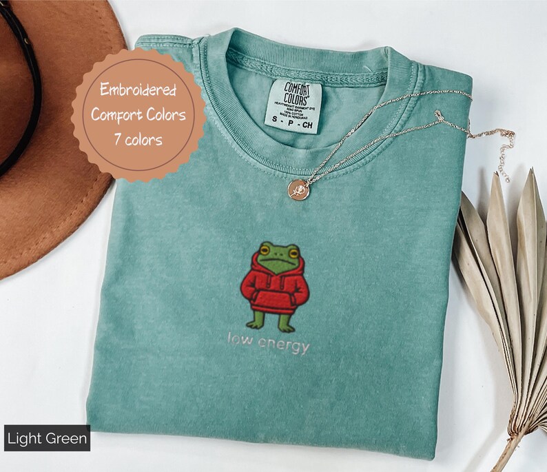May include: Light green t-shirt with an embroidered frog wearing a red hoodie. The text "low energy" is below the frog. The shirt has a Comfort Colors tag and a round tag that says "Embroidered Comfort Colors 7 colors".