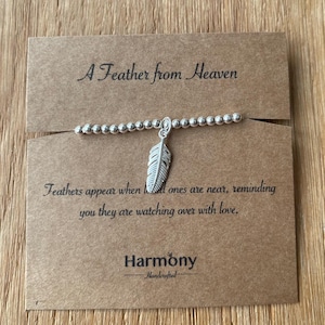 May include: A silver feather charm bracelet with a brown card that reads "A Feather from Heaven" and "Feathers appear when loved ones are near, reminding you they are watching over with love." The card also has the word "Harmony" and "Handcrafted" on it.