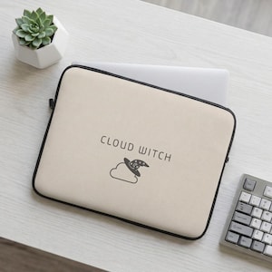 Puede incluir: Una funda para portátil de color beige con borde negro que presenta el texto CLOUD WITCH en una fuente sans-serif sobre un gráfico de líneas negras de una nube con un sombrero de mago con estrellas y un diseño de luna creciente en la copa.