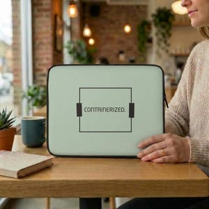Może przedstawiać: Jasnozielony pokrowiec na laptopa w kolorze szałwii z wyśrodkowaną czarną prostokątną grafiką i napisem "CONTAINERIZED." wykonanym czcionką bezszeryfową. Projekt zawiera dwa małe czarne pionowe paski po bokach prostokąta.