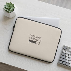 Pode incluir: Uma capa para portátil bege com rebordo preto, apresentando o texto "GOOD THINGS COMPILING" acima de um gráfico de barra de progresso parcialmente preenchido. A capa é retangular com cantos arredondados e fecho de correr.
