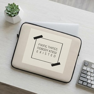 Puede incluir: Una funda para portátil de color beige con un gráfico de marco rectangular negro y dos tiras de cinta adhesiva negra en las esquinas. El texto dentro del marco dice: FIXING THINGS NOBODY KNEW EXISTED en una fuente negra sans-serif.