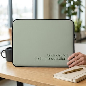 Könnte beinhalten: Eine salbeigrüne, rechteckige Laptoptasche mit schwarzem Rand und schwarzem Reißverschluss. Der Text "kinda chic to fix it in production" ist in Dunkelgrün auf der unteren rechten Seite aufgedruckt, ergänzt durch eine vertikale Linie.