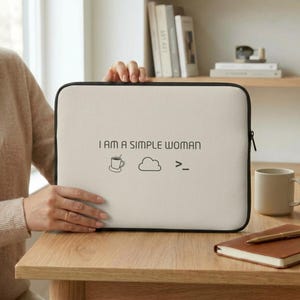 Op de afbeelding: Een rechthoekige beige laptophoes met zwarte rand en zwarte rits. De voorzijde is voorzien van de tekst I AM A SIMPLE WOMAN, gevolgd door een lijntekening van een koffiekopje, een wolk en een symbool voor een computeropdrachtprompt.