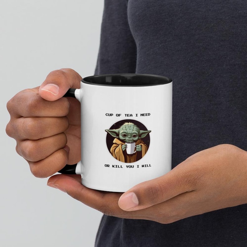 K&ouml;nnte beinhalten: Ein wei&szlig;er Keramikbecher mit schwarzem Innenraum und Henkel. Die Vorderseite zeigt eine kreisf&ouml;rmige Grafik eines gr&uuml;nen Wesens in einer braunen Robe, das eine kleine wei&szlig;e Tasse h&auml;lt. Dar&uuml;ber steht CUP OF TEA I NEED und darunter OR KILL YOU I WILL.