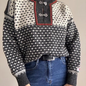 Könnte beinhalten: Ein anthrazitfarbener und cremefarbener Strickpullover mit wiederkehrendem geometrischem Muster, Rundhalsausschnitt mit zwei silberfarbenen Metallverschlüssen und roter Stickborte entlang der Knopfleiste. Der Pullover hat gerippte Bündchen.