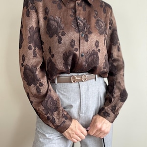 Pode incluir: Camisa marrom com estampa floral escura. A camisa de manga comprida tem gola e é combinada com calças cinza e um cinto marrom. O desenho floral é marrom escuro.