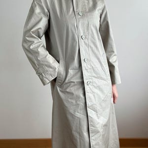 May include: A light beige trench coat with a classic design. The coat features a collared neckline, button-down front, and long sleeves. It has a relaxed fit and a knee-length hemline. The coat has two front pockets and a button detail on the sleeves.