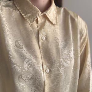 Op de afbeelding: Een lichtgeel overhemd met lange mouwen en een knoopsluiting, voorzien van een ton-sur-ton paisley-patroon over de gehele stof. Het overhemd heeft een klassieke kraag, een knoopsluiting aan de voorzijde met bijpassende lichte knopen en een gladde, licht glanzende afwerking.