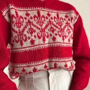 May include: A red v-neck sweater with a cream geometric pattern across the chest and upper sleeves. The sweater has long sleeves and a ribbed hem. The pattern features stylized floral and geometric shapes. The sweater is made of a knitted material.