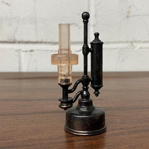 May include: A small, black metal lamp with a clear, cylindrical shade and a round base. The lamp features a detailed design with a vertical post and a bulbous reservoir. The shade is a light amber colour. The lamp is on a wooden surface.