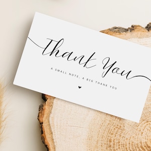 May include: A white card with the words "Thank You" in elegant black script. Below, smaller text reads "A small note, a big thank you," with a small heart. The card rests on a wooden surface.