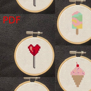 Op de afbeelding: Vijf borduurringen met verschillende kruissteekontwerpen van snoep en zoetigheden. De ontwerpen omvatten een groene lolly, een roze en groene ijslolly, een rode hartlolly, een roze cupcake met sprankelingen en een groen en geel ingepakt snoepje.