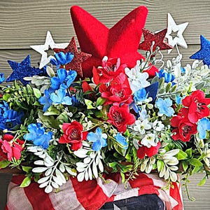 May include: A patriotic floral arrangement featuring red, white, and blue flowers and star-shaped decorations. The arrangement includes red roses, blue and white blossoms, and decorative stars, all set against a wooden background.
