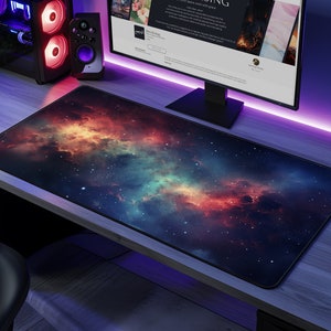 Op de afbeelding: Een grote, kleurrijke muismat met een galaxie-thema, met een donkerblauwe achtergrond en wervelende rode, oranje en groene nevels. De muismat ligt op een houten bureau met een computermonitor en andere computerapparatuur.