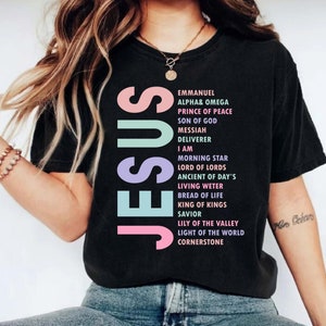 May include: Black t-shirt with the word "JESUS" in pastel colors. Below the word are the words "EMMANUEL", "ALPHA & OMEGA", "PRINCE OF PEACE", "SON OF GOD", "MESSIAH", "DELIVERER", "I AM", "MORNING STAR", "LORD OF LORDS", "ANCIENT OF DAY'S", "LIVING WETER", "BREAD OF LIFE", "KING OF KINGS", "SAVIOR", "LILY OF THE VALLEY", "LIGHT OF THE WORLD", and "CORNERSTONE".