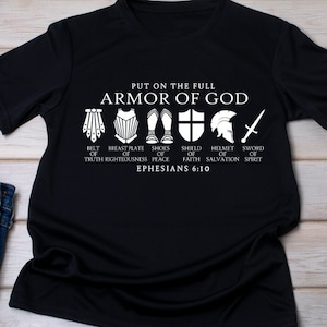 May include: Black t-shirt with a white graphic of the Armor of God. The graphic includes a belt, breastplate, shoes, shield, helmet, and sword. The text "PUT ON THE FULL ARMOR OF GOD" is at the top of the graphic. The text "BELT OF TRUTH, BREASTPLATE OF RIGHTEOUSNESS, SHOES OF PEACE, SHIELD OF FAITH, HELMET OF SALVATION, SWORD OF SPIRIT" is below the graphic. The text "EPHESIANS 6:10" is at the bottom of the graphic.