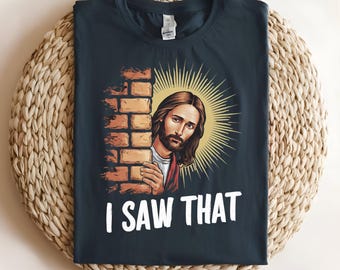 May include: A dark gray t-shirt with a graphic of Jesus peeking around a brick wall. The text "I SAW THAT" is printed below the image.