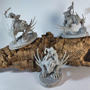 Può includere: Tre figure in miniatura stampate in 3D di colore grigio di una creatura con spine affilate e un cavaliere sulla sua schiena. Le figure sono posizionate su una superficie di legno marrone.