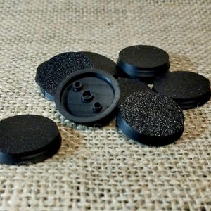 Könnte beinhalten: Eine Ansammlung schwarzer, kreisförmiger Sockel mit strukturierten Oberflächen. Einige Sockel haben einen glatten Rand, während andere eine strukturierte Oberfläche aufweisen. Ein Sockel hat drei Löcher. Die Sockel sind wahrscheinlich für Miniaturen oder Modelle.