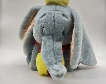 May include: A gray plush toy elephant with large ears, wearing a red and yellow hat. The elephant is holding a yellow ball in its trunk.