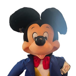 Peut inclure: Une peluche de Mickey Mouse portant un costume bleu avec un n&oelig;ud papillon rouge et un gilet jaune &agrave; pois. La peluche a des oreilles noires et un nez brun.