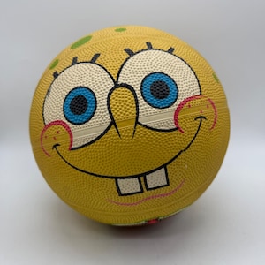 Puede incluir: Un balón de baloncesto amarillo con el diseño de una cara de personaje de dibujos animados, con grandes ojos blancos, iris azul brillante, pestañas negras, una boca sonriente con dos dientes frontales y mejillas rosadas sobre goma texturizada.