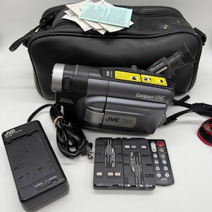 Könnte beinhalten: Schwarzer und grauer JVC Compact VHS Camcorder mit Zubehör, darunter ein Ladegerät, Batterien und eine Fernbedienung. Der Camcorder hat ein silbernes Objektiv und den Text "100x" an der Seite. Eine schwarze Tragetasche befindet sich im Hintergrund.